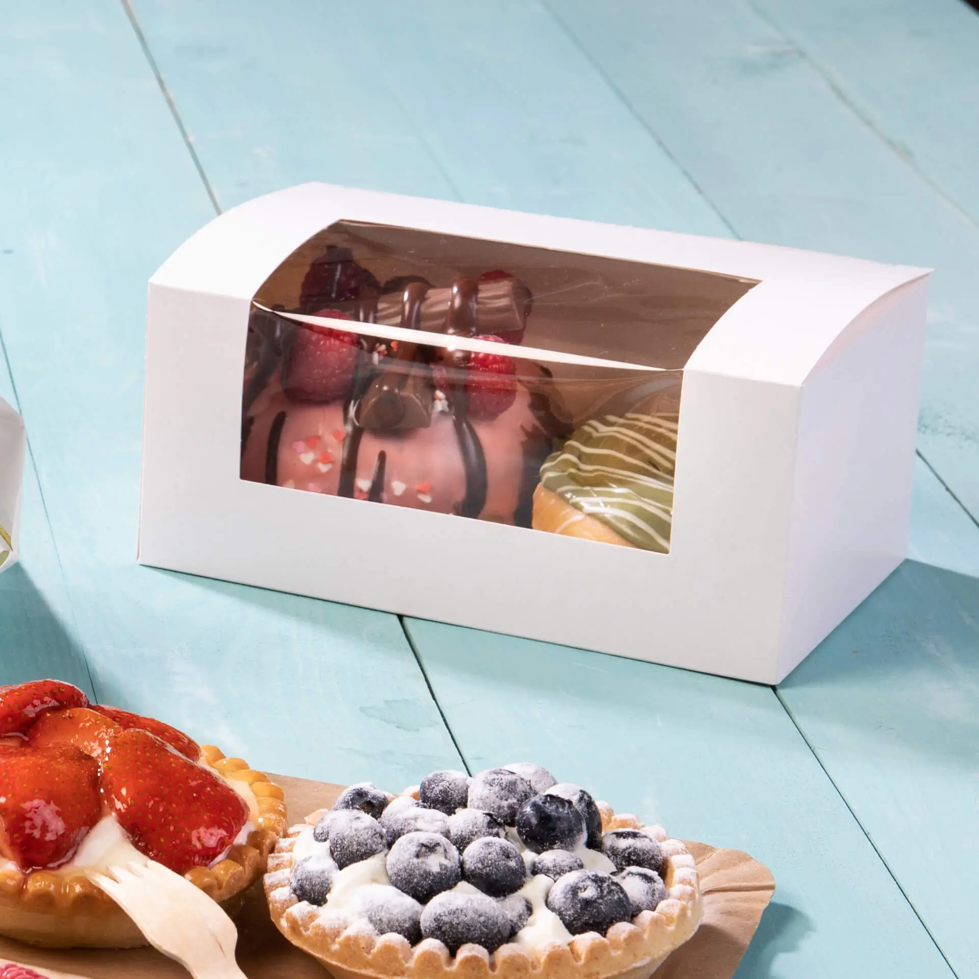 Weiße Cupcake-Box mit Cellulosefenster, die eine Auswahl an Gebäck zeigt, darunter Kuchen und Torten, auf einem türkisfarbenen Untergrund.