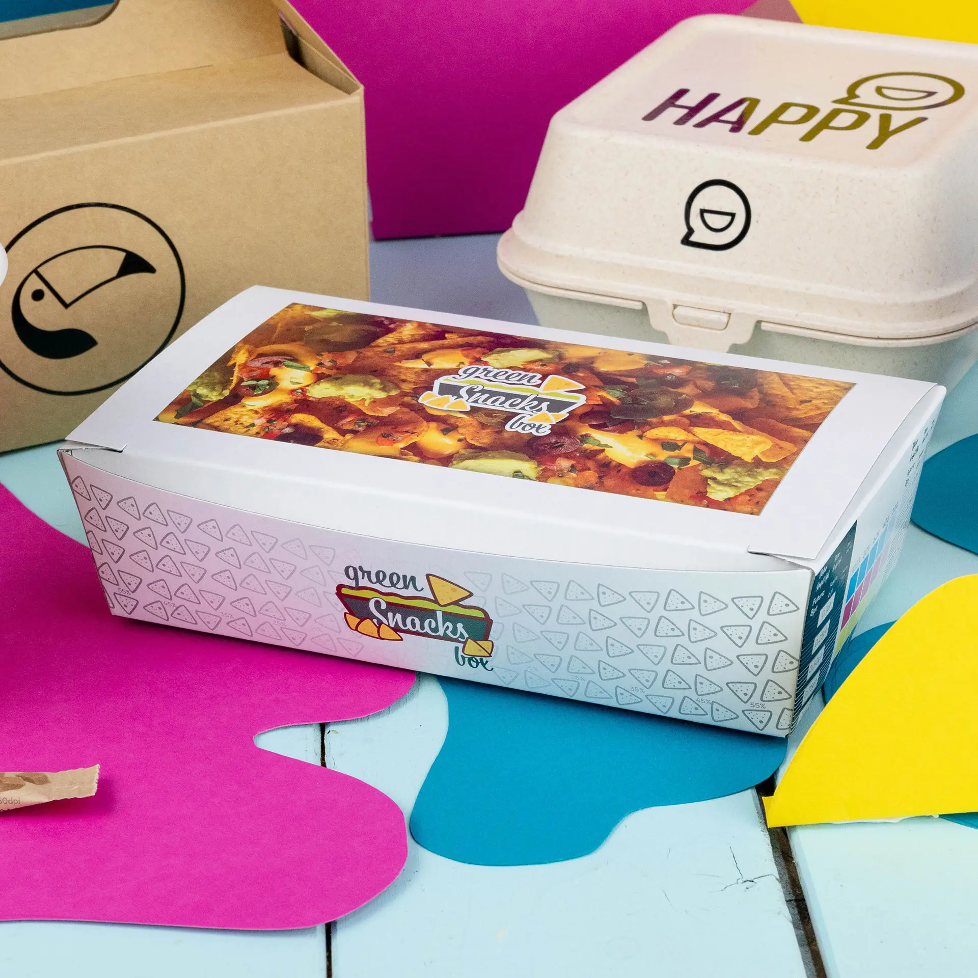 Boîte à snacks blanche avec un design imprimé coloré, étiquetée pour les snacks, sur un fond vibrant avec un emballage similaire à proximité.