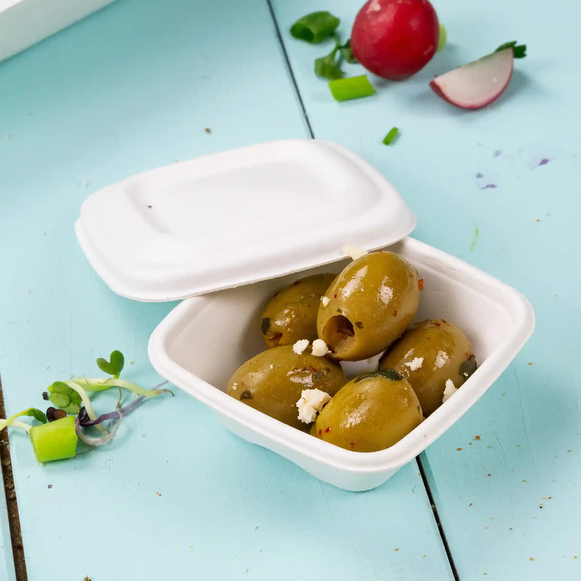 Bicchiere quadrato bianco contenente diverse olive verdi, posato su una superficie azzurra con verdure sparse.