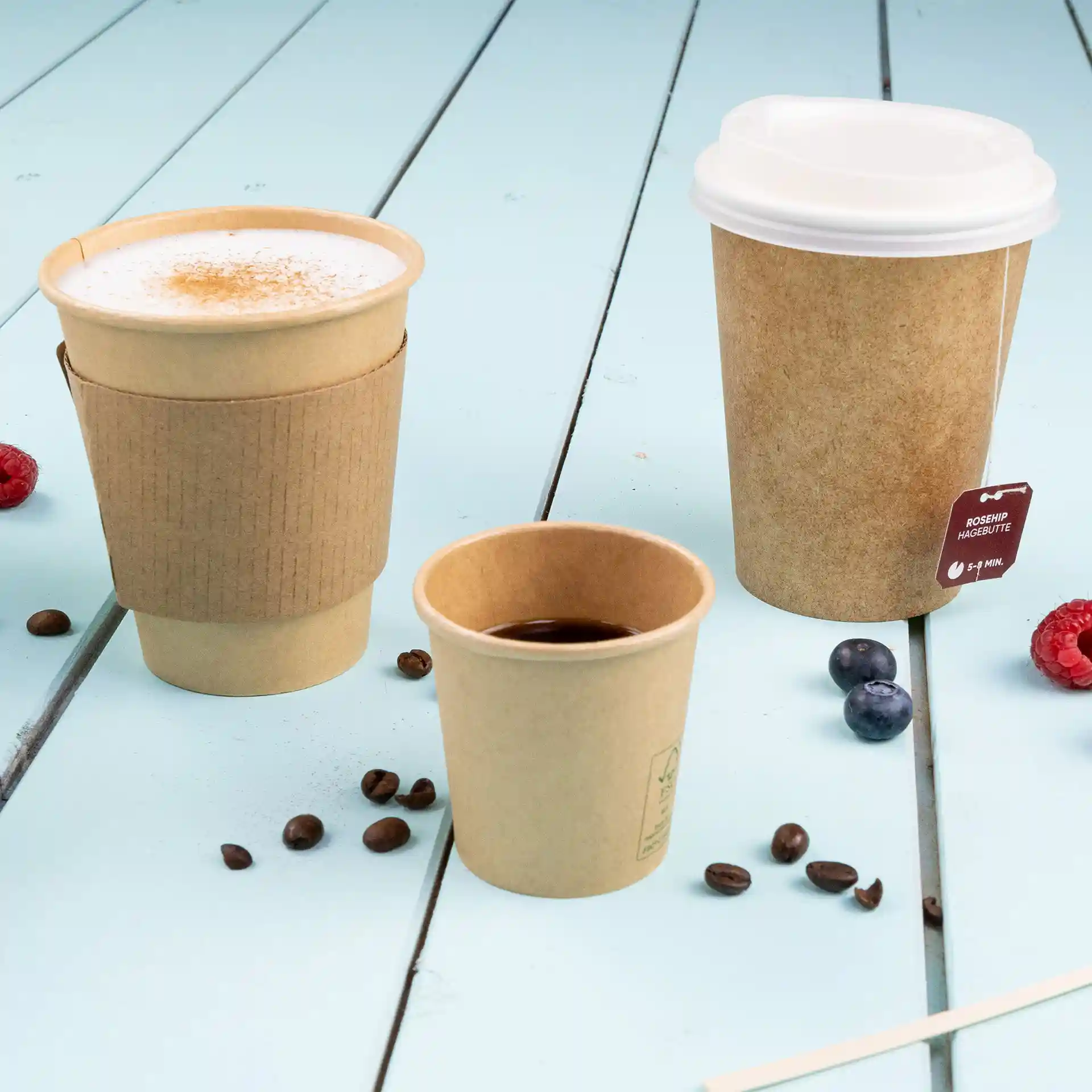 Image de trois gobelets en papier brun de tailles variées sur une surface en bois bleu, avec des grains de café et des baies éparpillés.