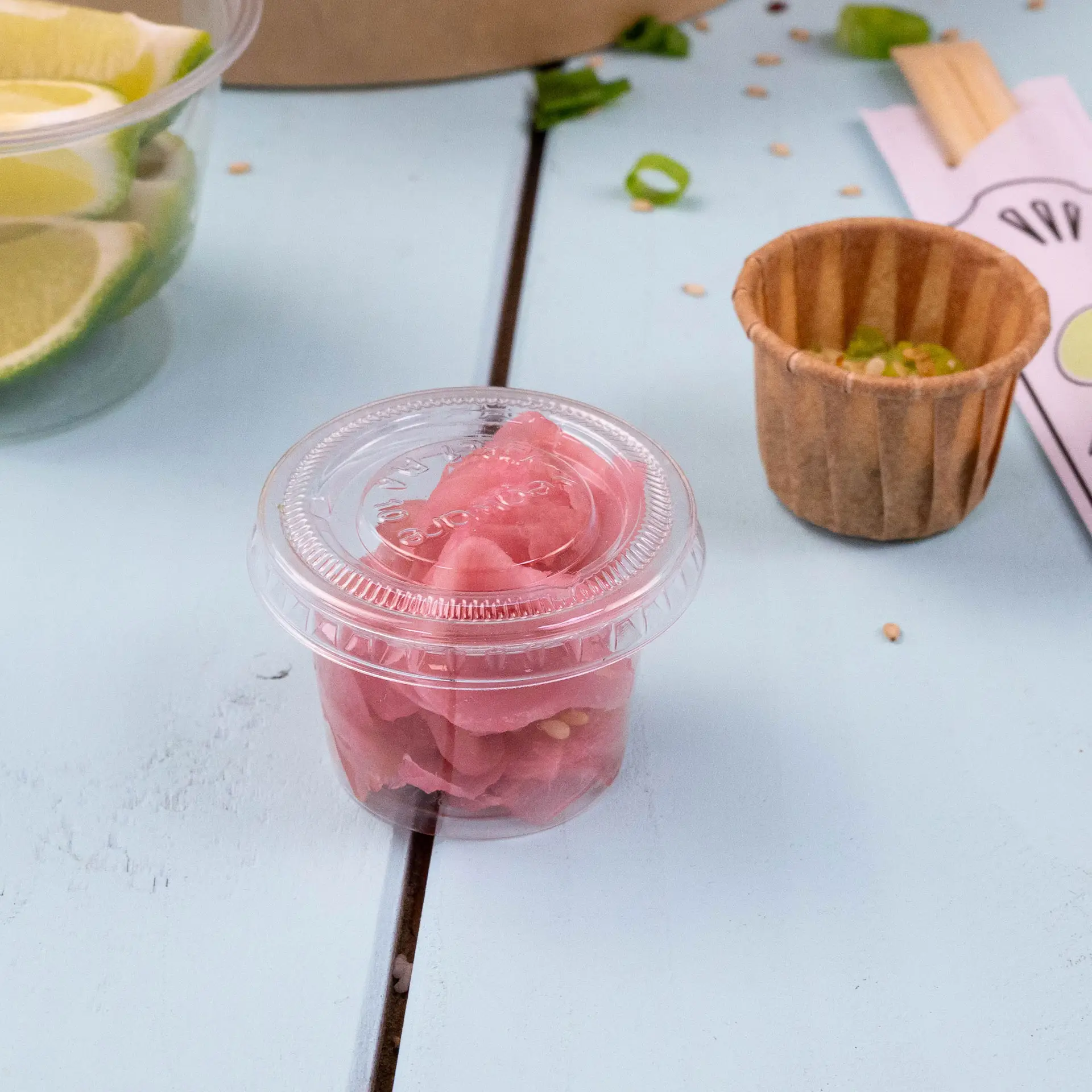 Un gobelet en plastique transparent contenant du gingembre mariné rose sur une surface bleu clair, avec un gobelet en papier brun et des quartiers de citron vert en arrière-plan.