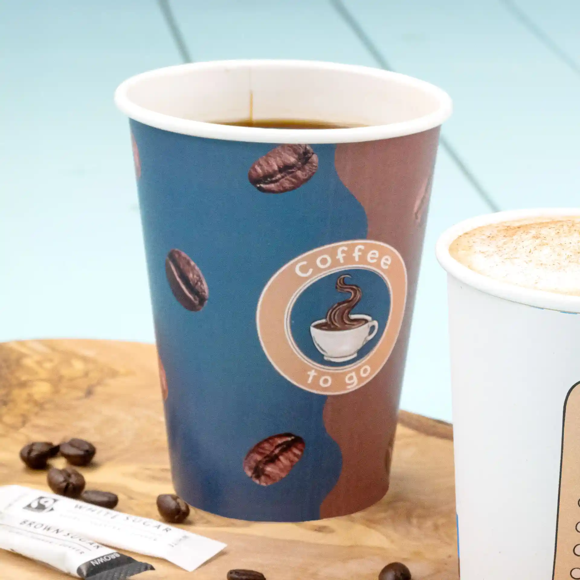 Bicchiere di carta con design stampato, riempito di caffè, posato su una superficie di legno con bustine di zucchero e chicchi di caffè.