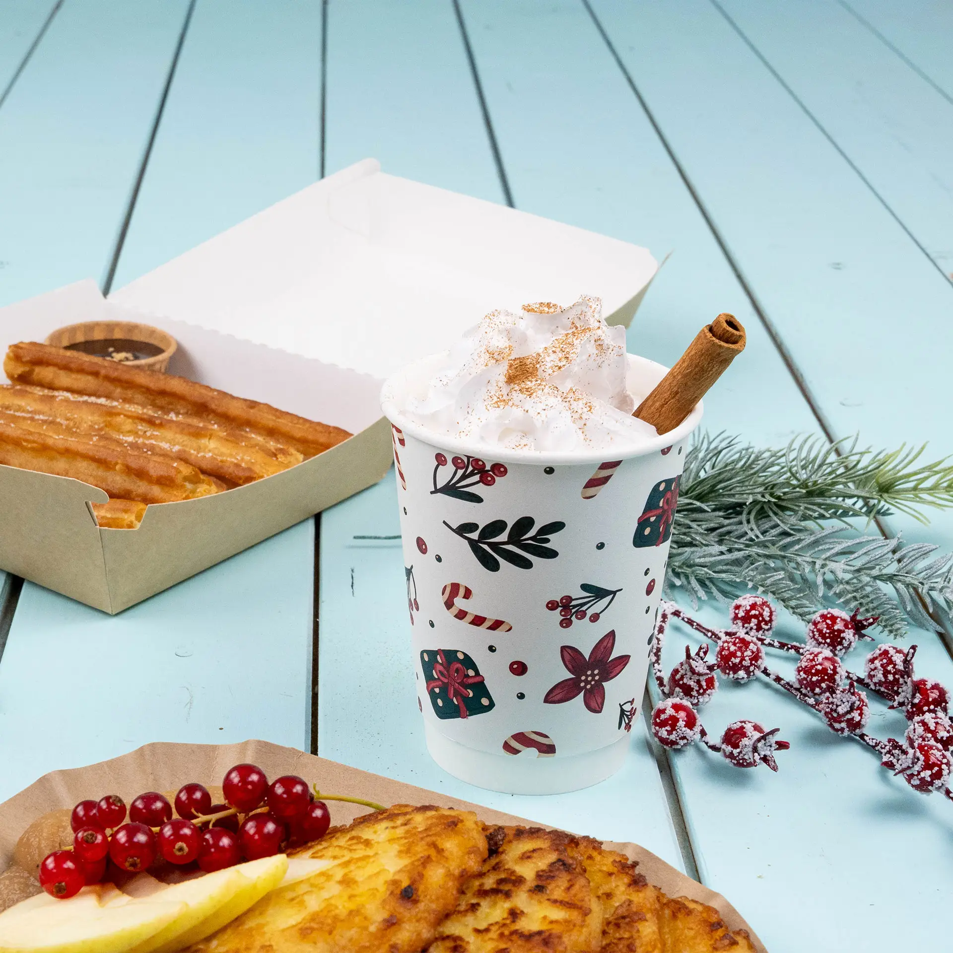 Doppelwandiger Becher mit Winterdesign, gefüllt mit Sahne und einem Zimtstock, neben Churros und Obst.