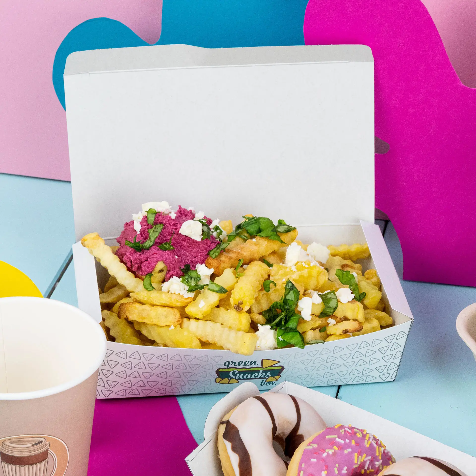 Une boîte à snacks blanche remplie de frites ondulées, garnie d'une tartinade rose, de feta et d'herbes, sur des fonds colorés.