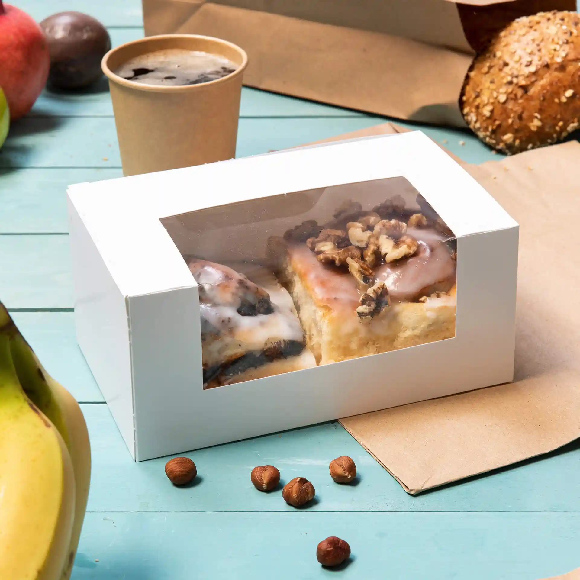 Weiße Pappschachtel mit Zellulosefenster, die zwei Gebäckstücke enthält, umgeben von einer Banane, Haselnüssen und einem Becher.