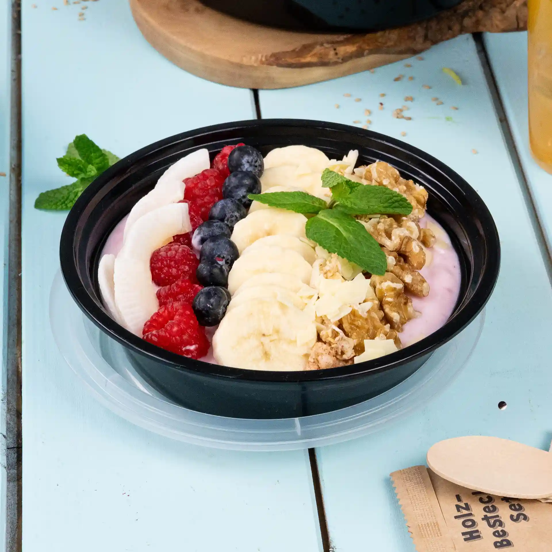 Piatto rotondo nero con yogurt, fette di banana, mirtilli, lamponi, granola e foglie di menta, su una superficie blu chiaro.