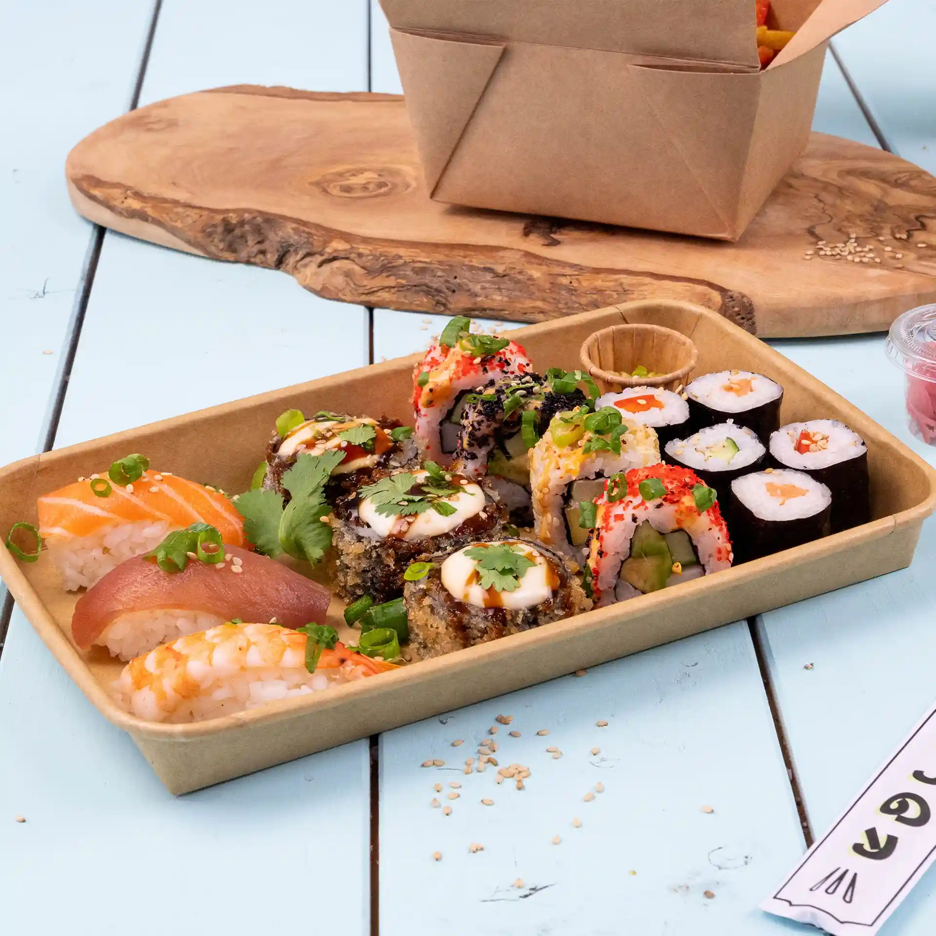 Sushi-Auswahl in einer braunen PE-beschichteten Box, mit verschiedenen Rollen und Sashimi auf einer blauen Holzoberfläche.