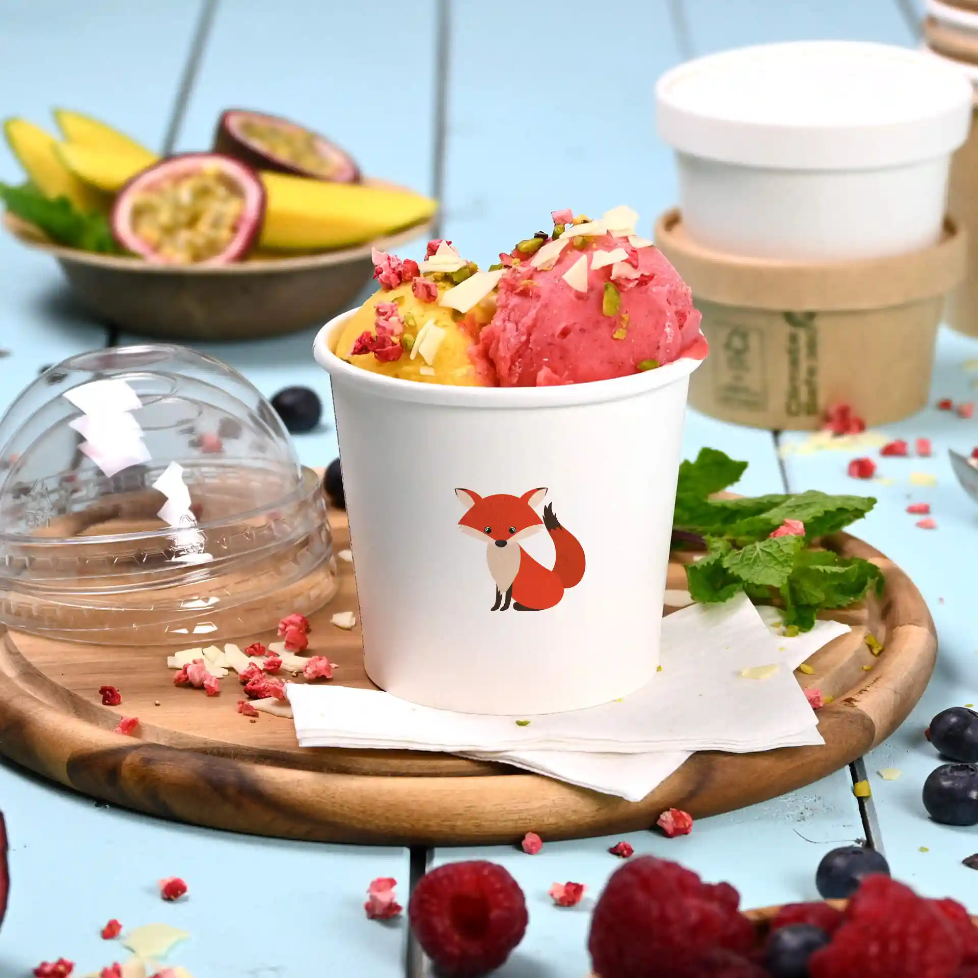 Runder weißer Becher mit Fuchsillustration, gefüllt mit pinkem und gelbem Eis, auf einem Holzuntersetzer.