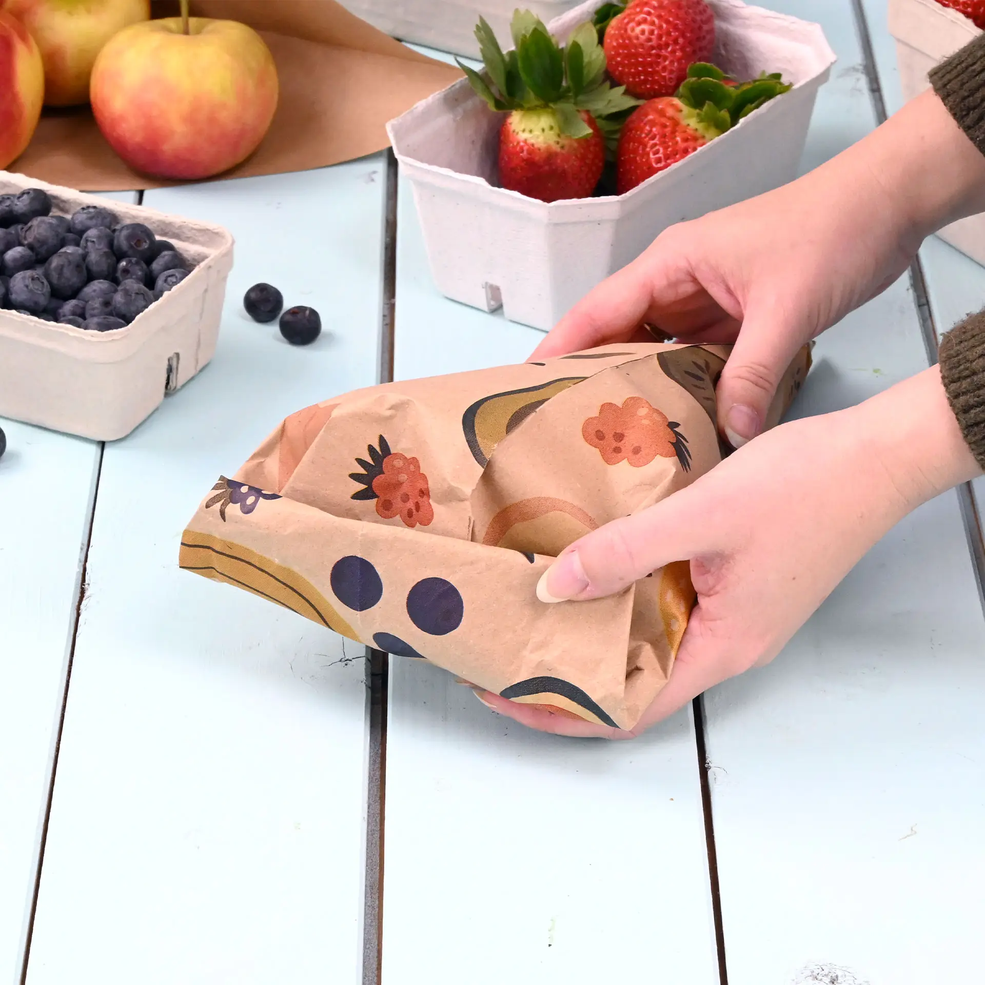 Hände halten einen bedruckten Papiercone mit einem braunen Design, umgeben von Obstkörben auf einem Holztisch.