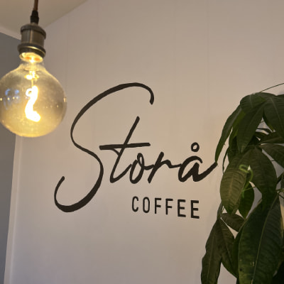 Logo sur le mur indiquant "Stora Coffee"; ampoule suspendue à proximité; plante verte au premier plan.