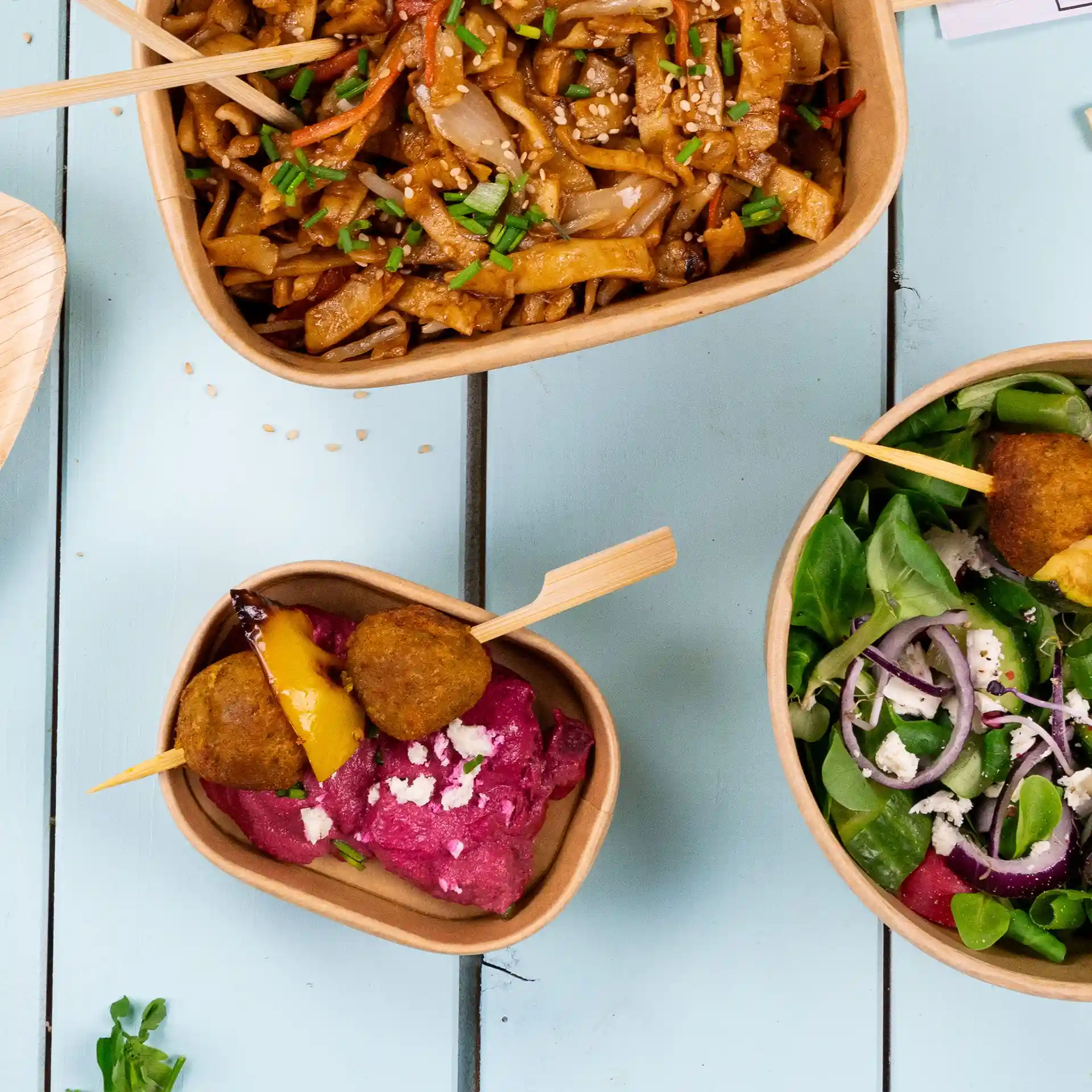 Immagine di tre contenitori alimentari rettangolari in kraft con vari piatti, tra cui noodles, insalata e una salsa rosa con spiedini.