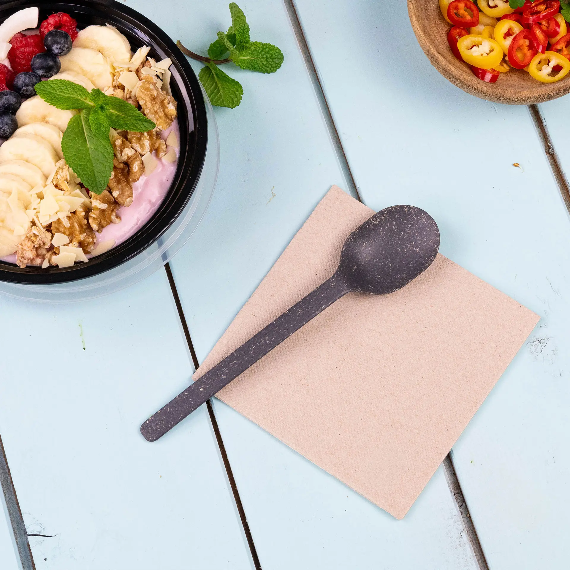 Bild eines schwarzen Löffels auf einer beigen Serviette, mit einer Schüssel Joghurt, garniert mit Früchten und Nüssen im Hintergrund.
