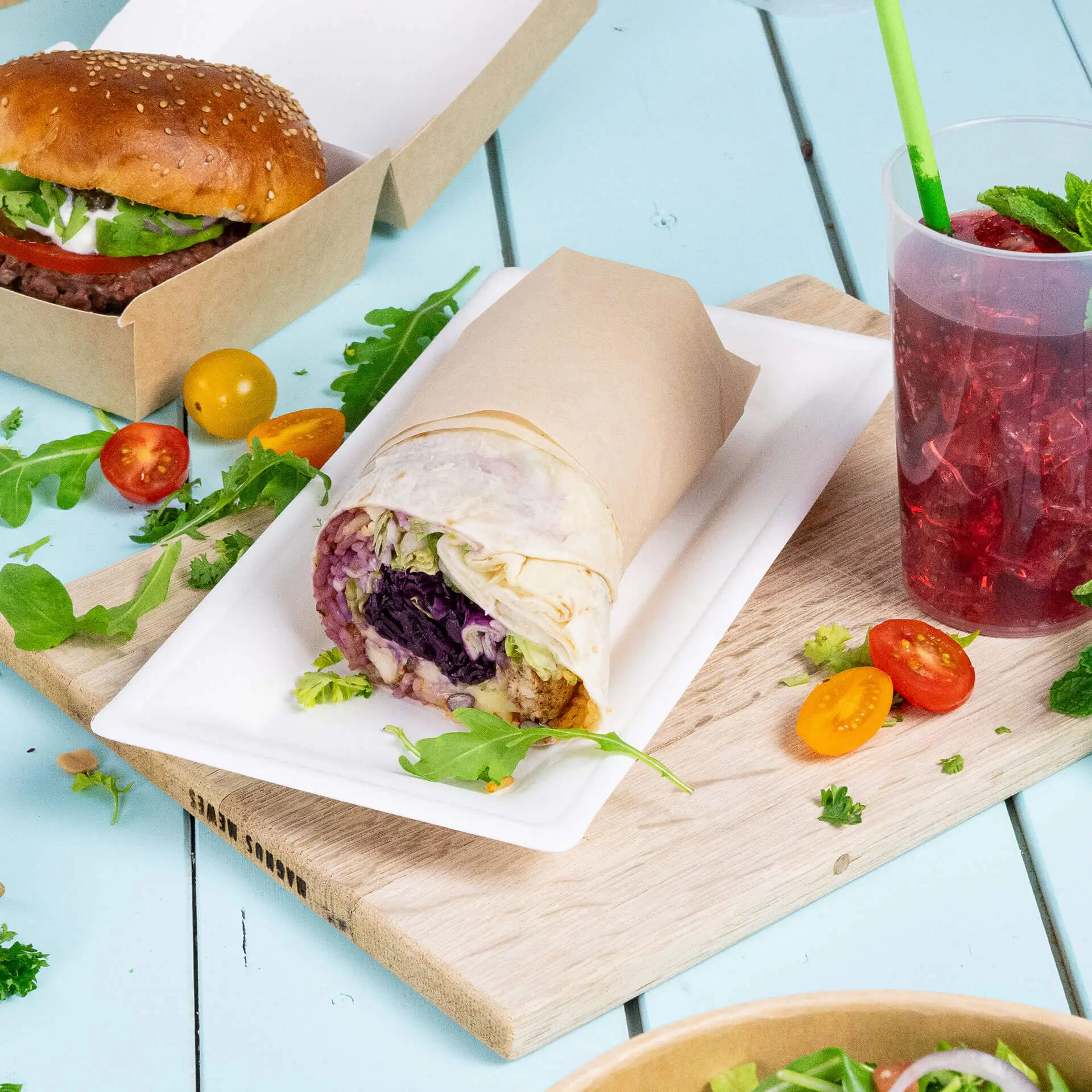 Piatto con un wrap su un piatto rettangolare in canna da zucchero, con un hamburger, bevanda e verdure varie sullo sfondo.