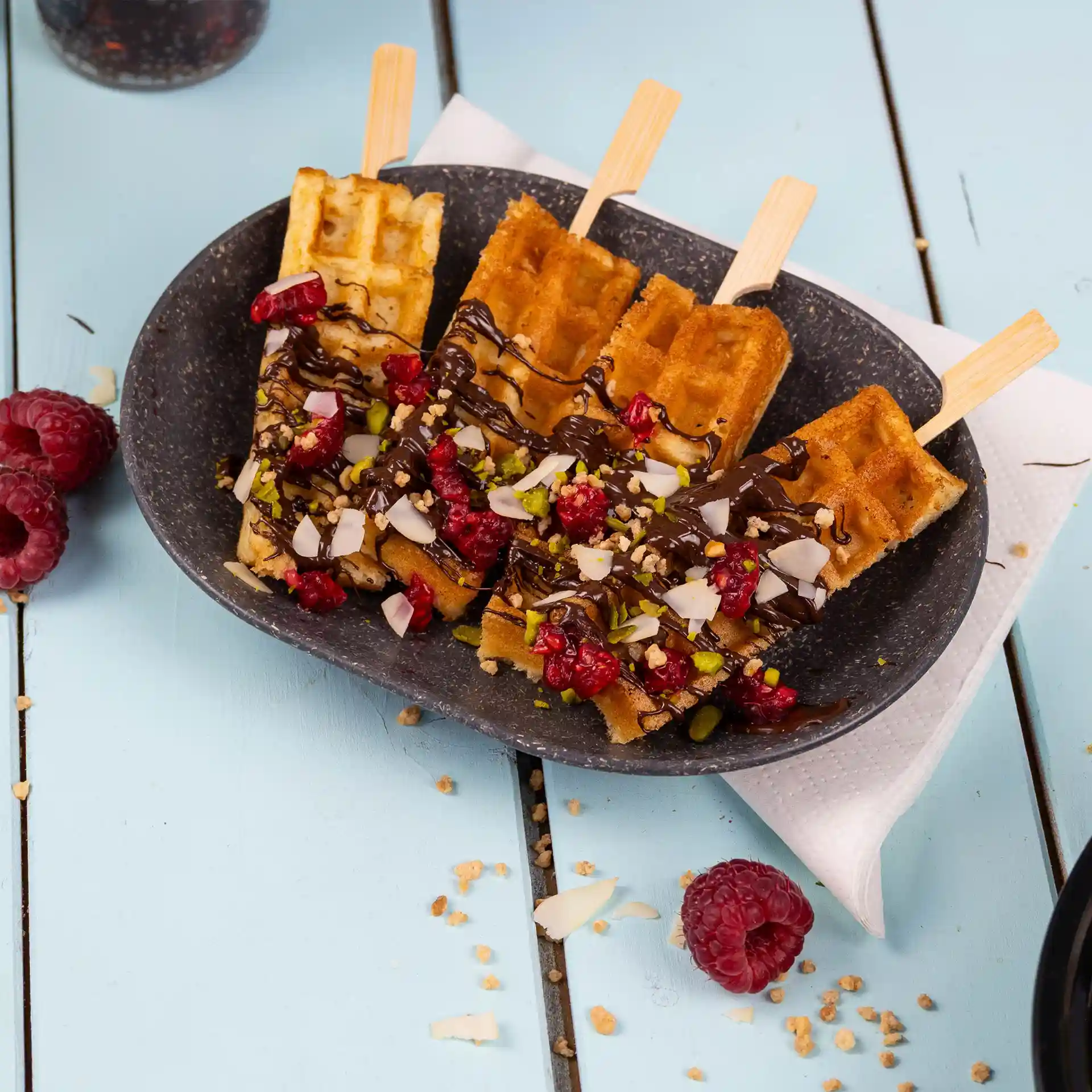 Assiette avec trois bâtonnets de gaufre garnis de chocolat, framboises et noix, sur un plat ovale gris sur fond bleu.