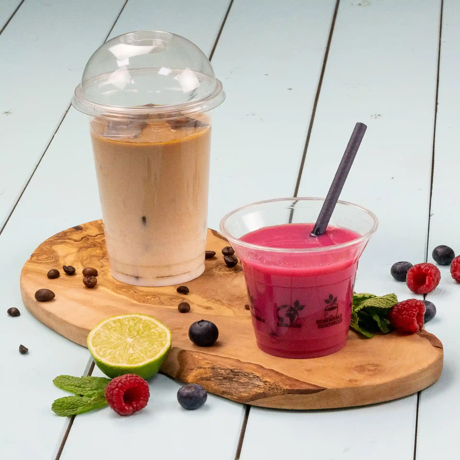 Immagine di un bicchiere trasparente con un coperchio a cupola contenente una bevanda marrone chiaro, accanto a una bevanda rosa in un bicchiere trasparente con una cannuccia, su un tagliere di legno con lime, frutti di bosco e menta.