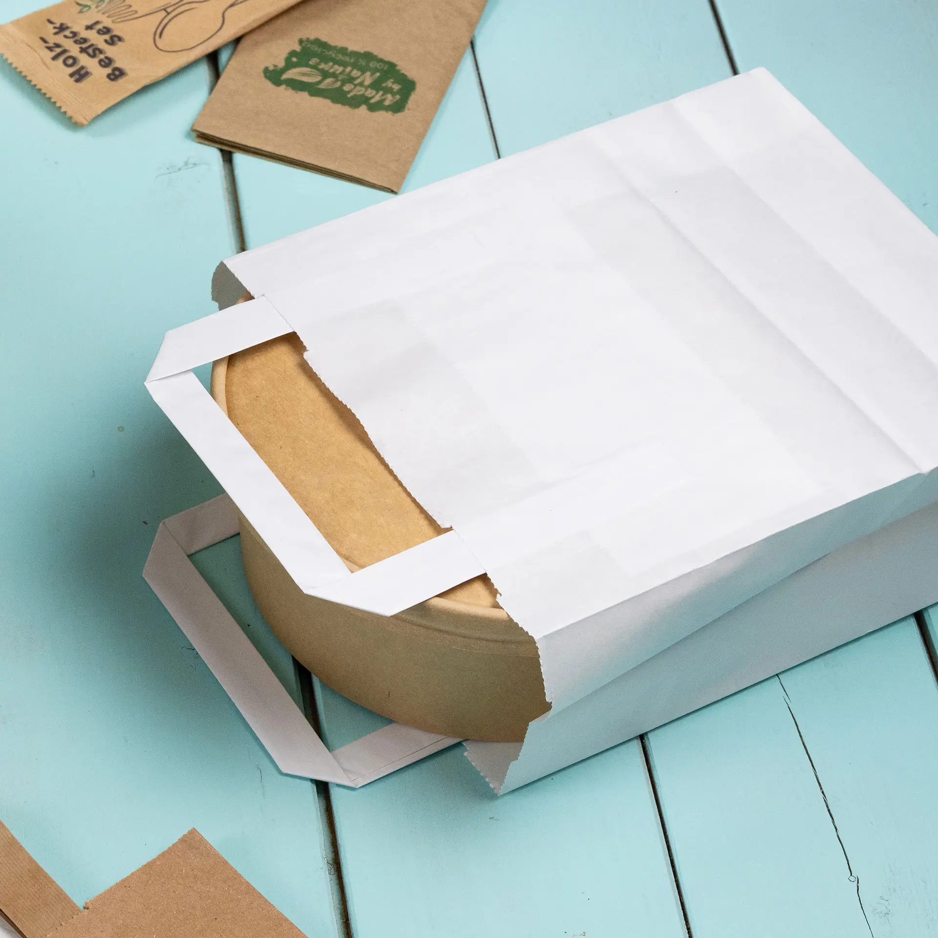 Weiße Papiertragetasche teilweise geöffnet, enthüllt einen runden Kraftpapierbehälter auf einer hellblauen Oberfläche.