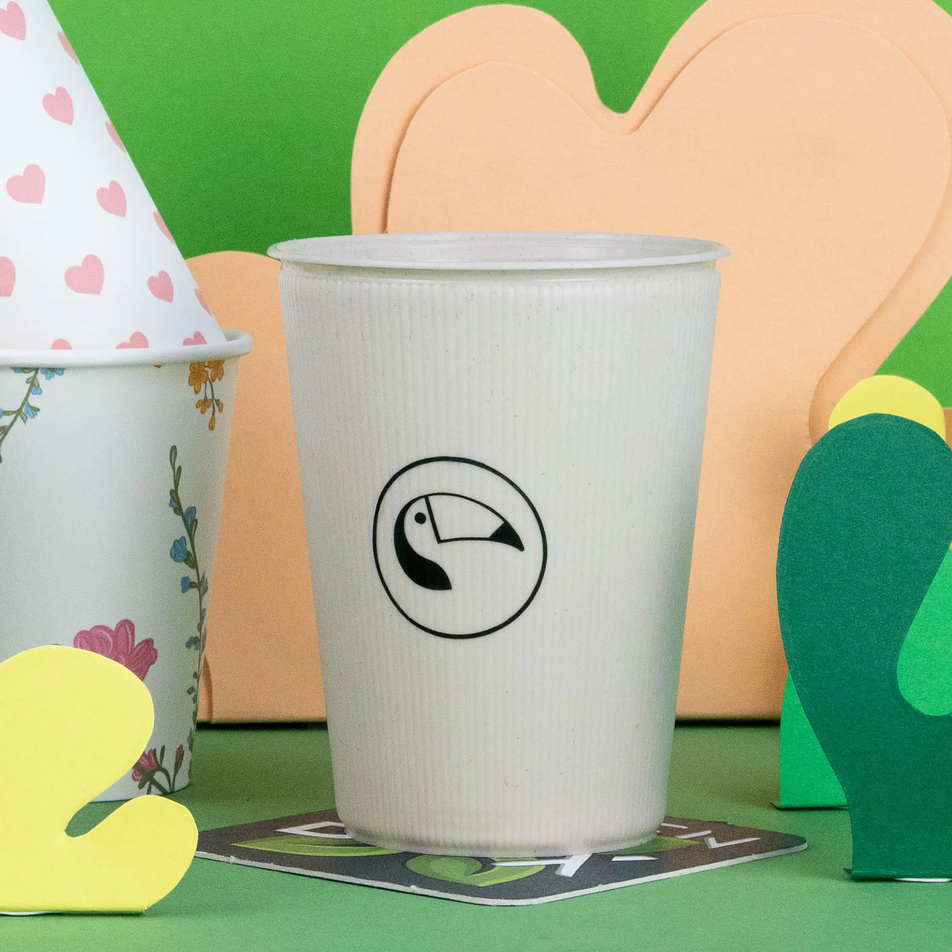 Gobelet réutilisable blanc à motifs ondulés avec un logo de toucan, placé parmi des décorations en papier colorées sur un fond vert.