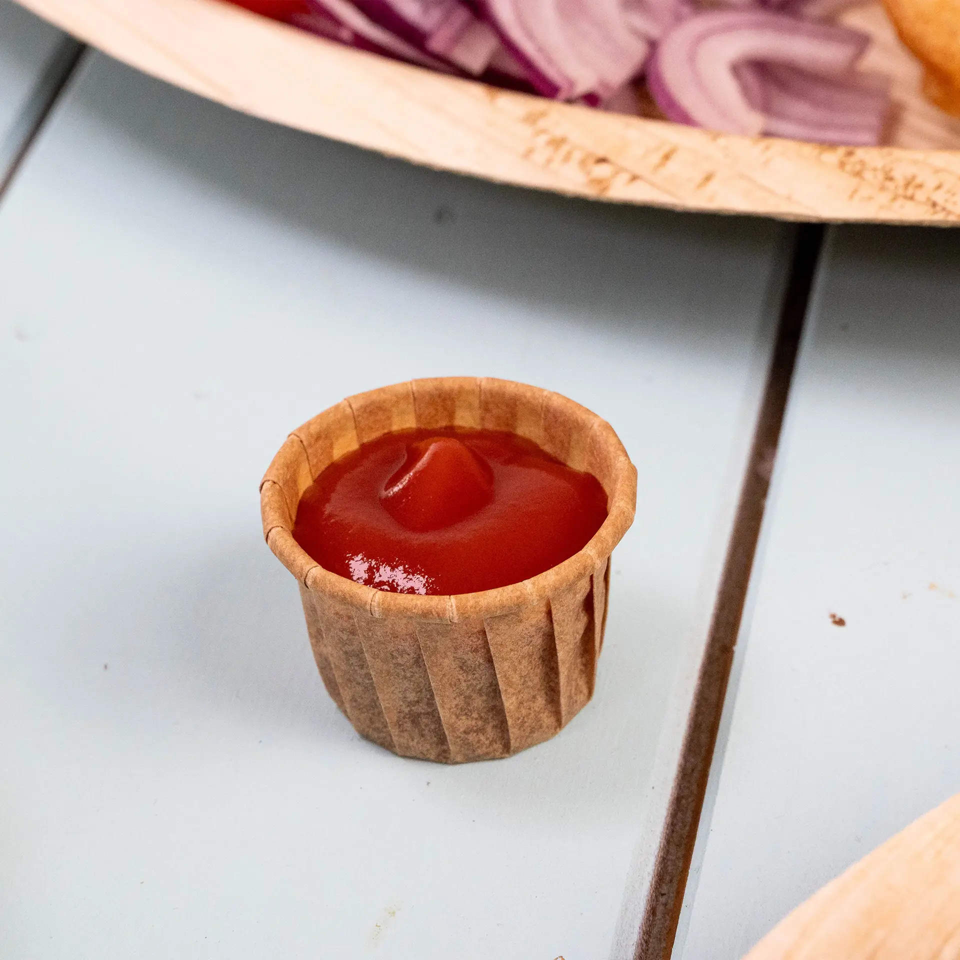 Coppetta di carta marrone piena di ketchup su una superficie azzurro chiaro, con cipolle rosse sfocate sullo sfondo.