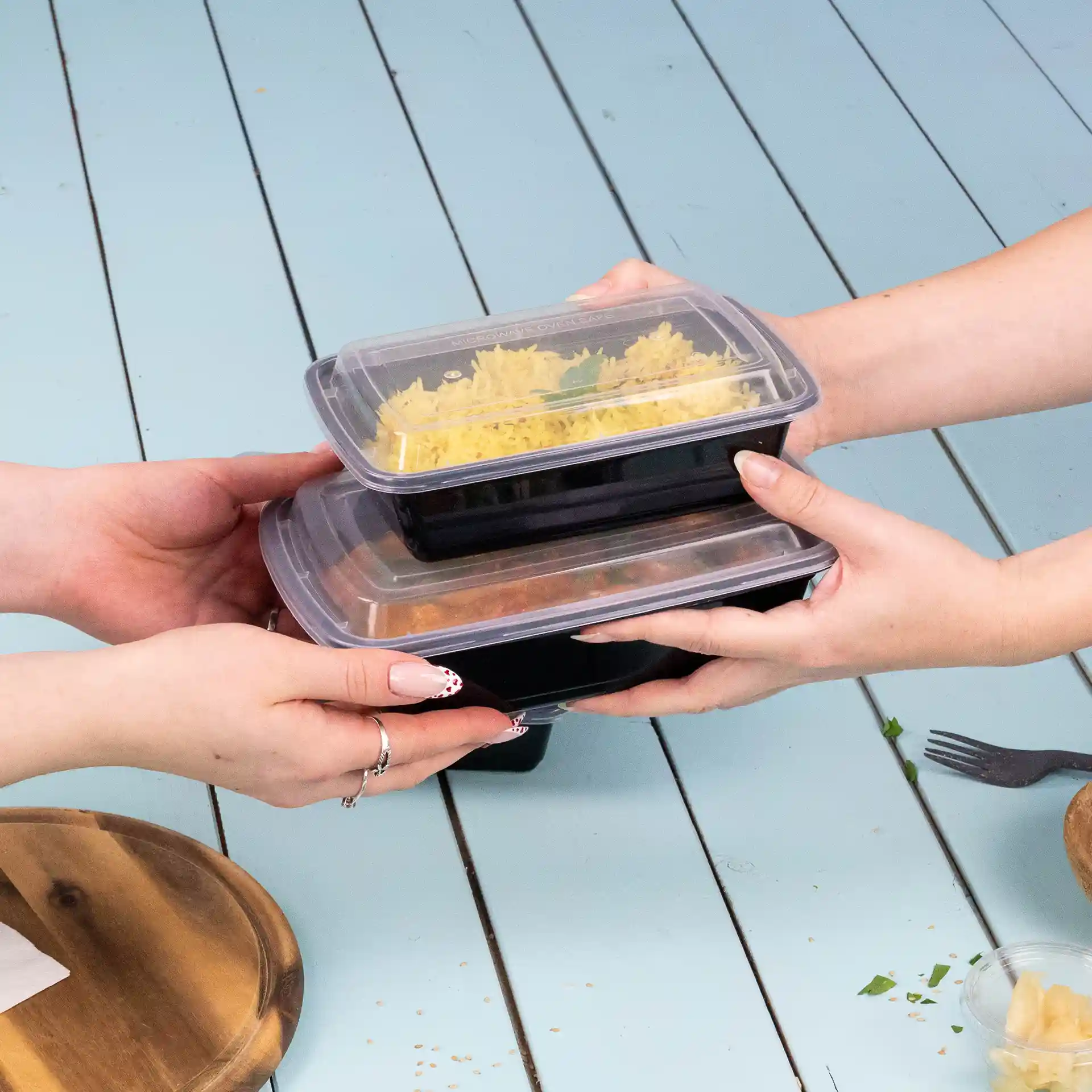 Due mani tengono contenitori alimentari rettangolari impilati con coperchi trasparenti, su una superficie blu chiaro.