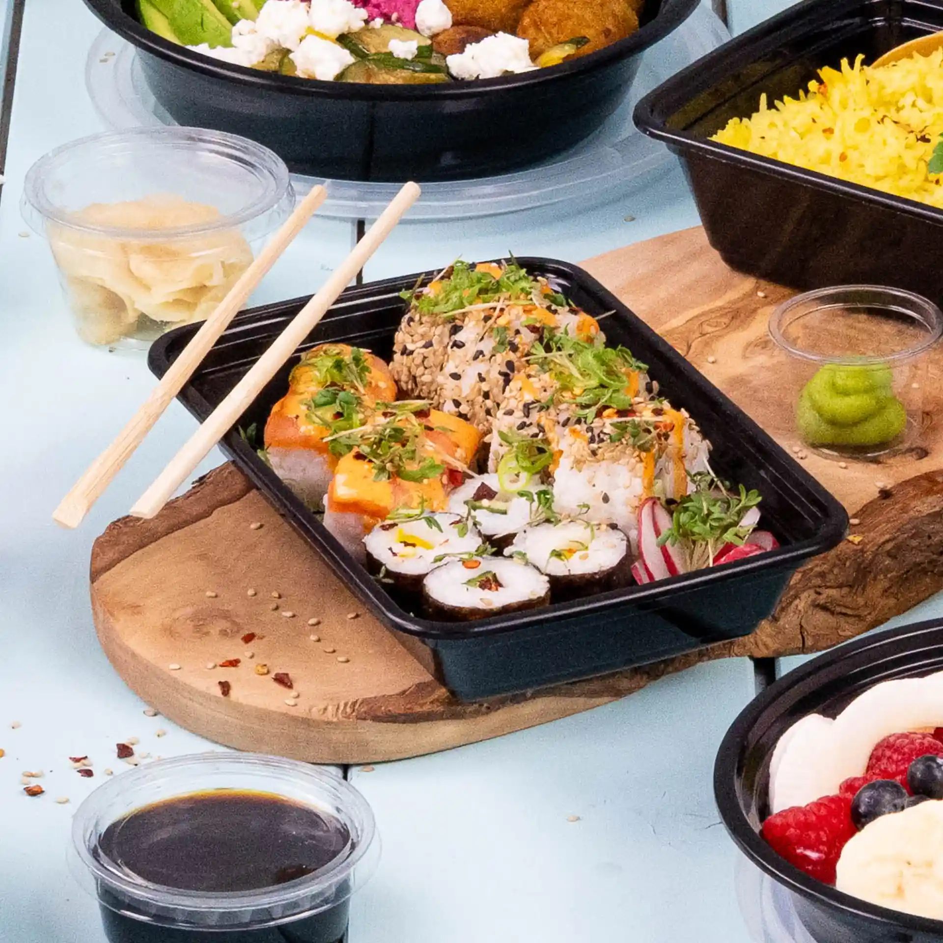 Sushi-Rollen mit Belag in einem schwarzen rechteckigen wiederverwendbaren Behälter, begleitet von Essstäbchen und Gewürzen.
