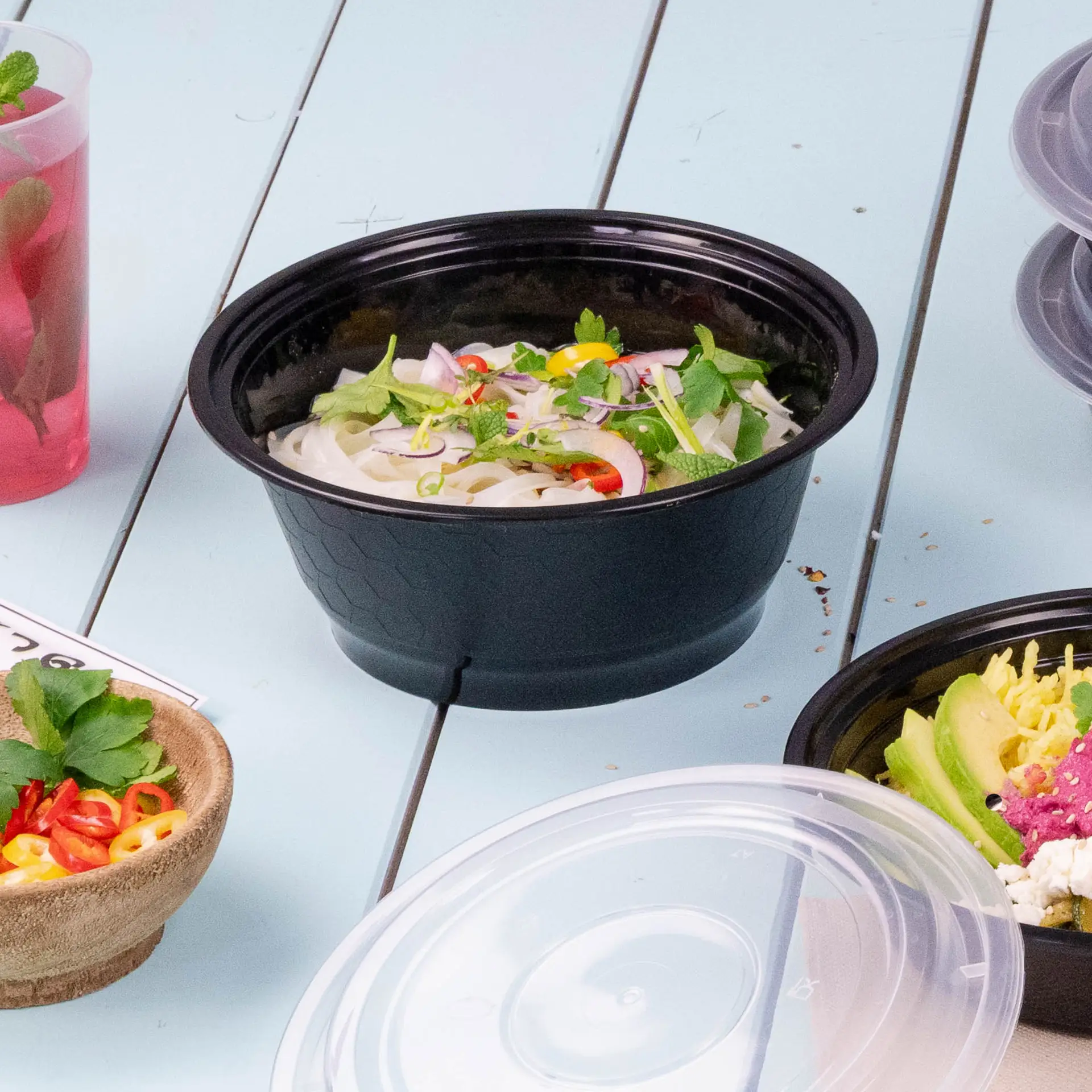 Ein schwarzer wiederverwendbarer Lebensmittelbehälter mit Nudeln, garniert mit Kräutern und Chili, umgeben von anderen Lebensmitteln.