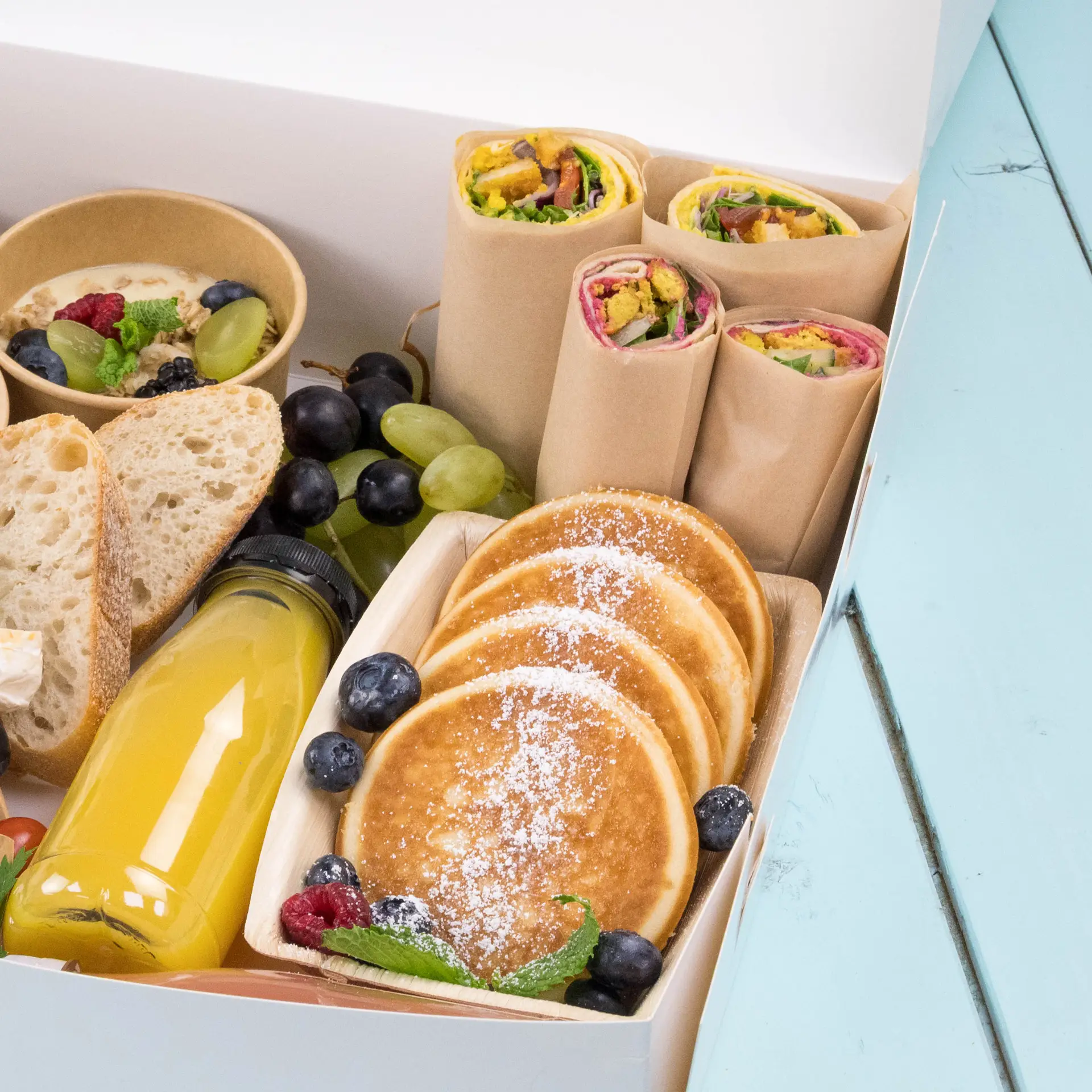 Scatola contenente pancake, wrap, frutta, pane e una bottiglia di succo, disposti per un pasto.
