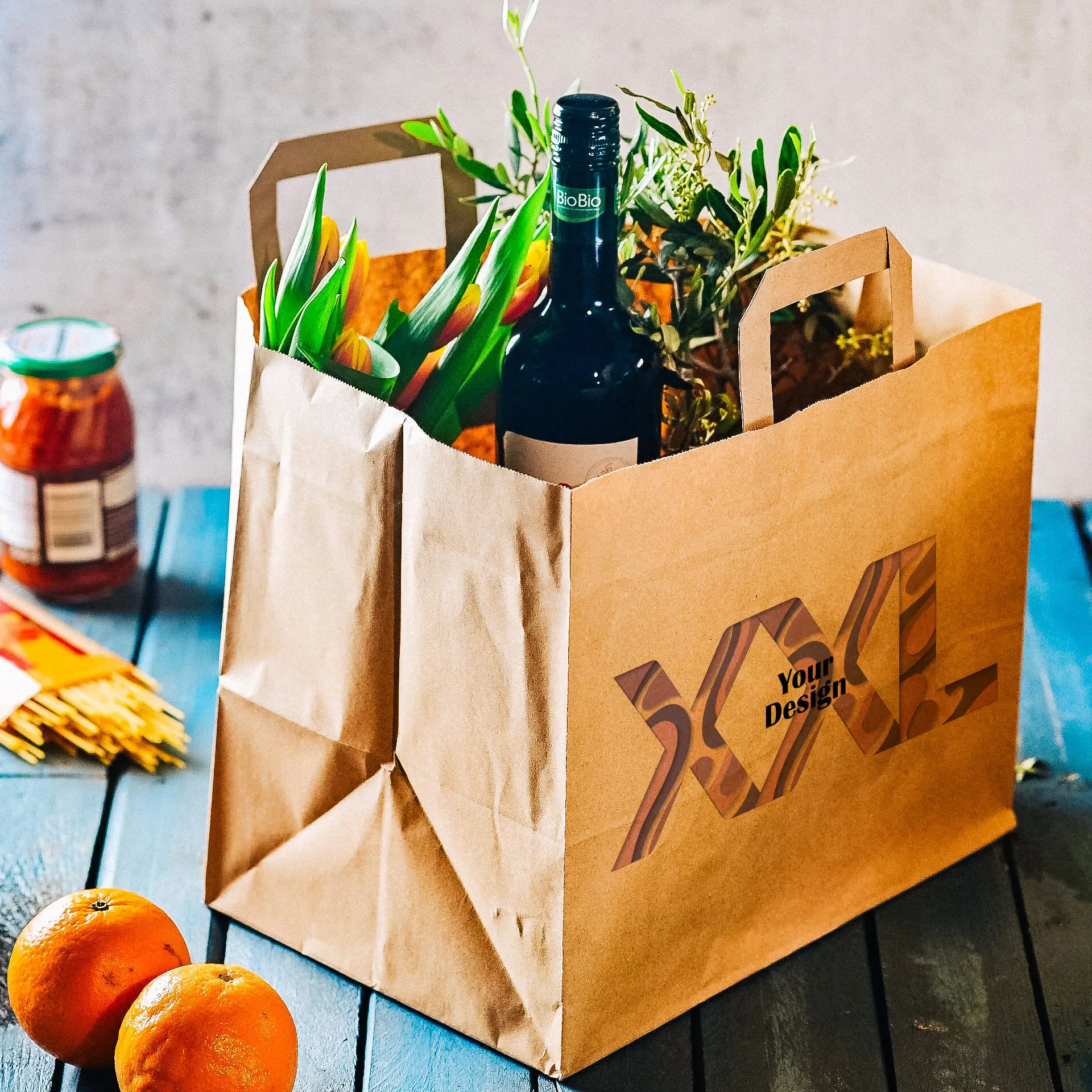 Borsa in carta personalizzabile realizzata in carta riciclata; contiene fiori, una bottiglia di vino e arance.