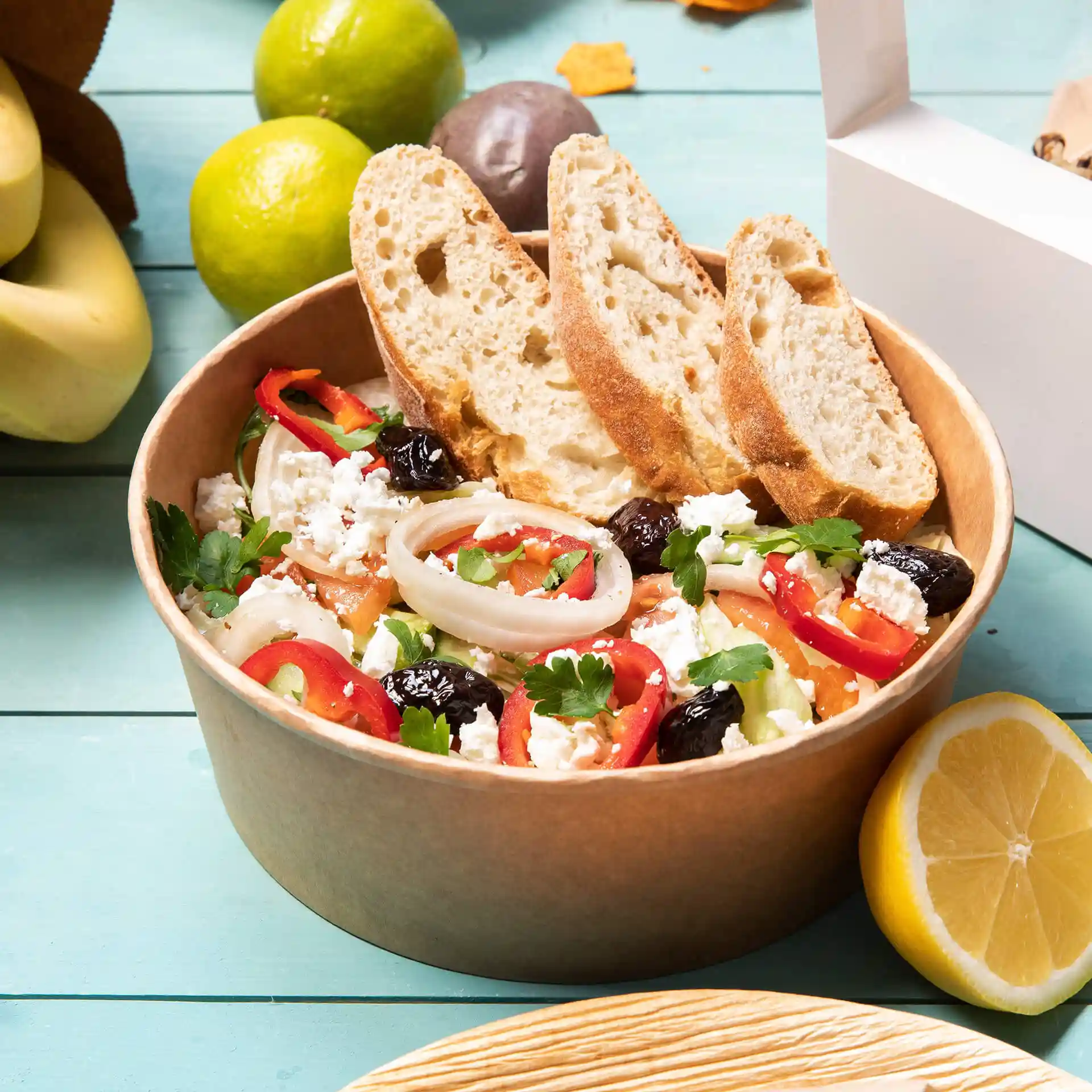 Ciotola rotonda di carta marrone piena di insalata, guarnita con pane, limone e varie verdure.