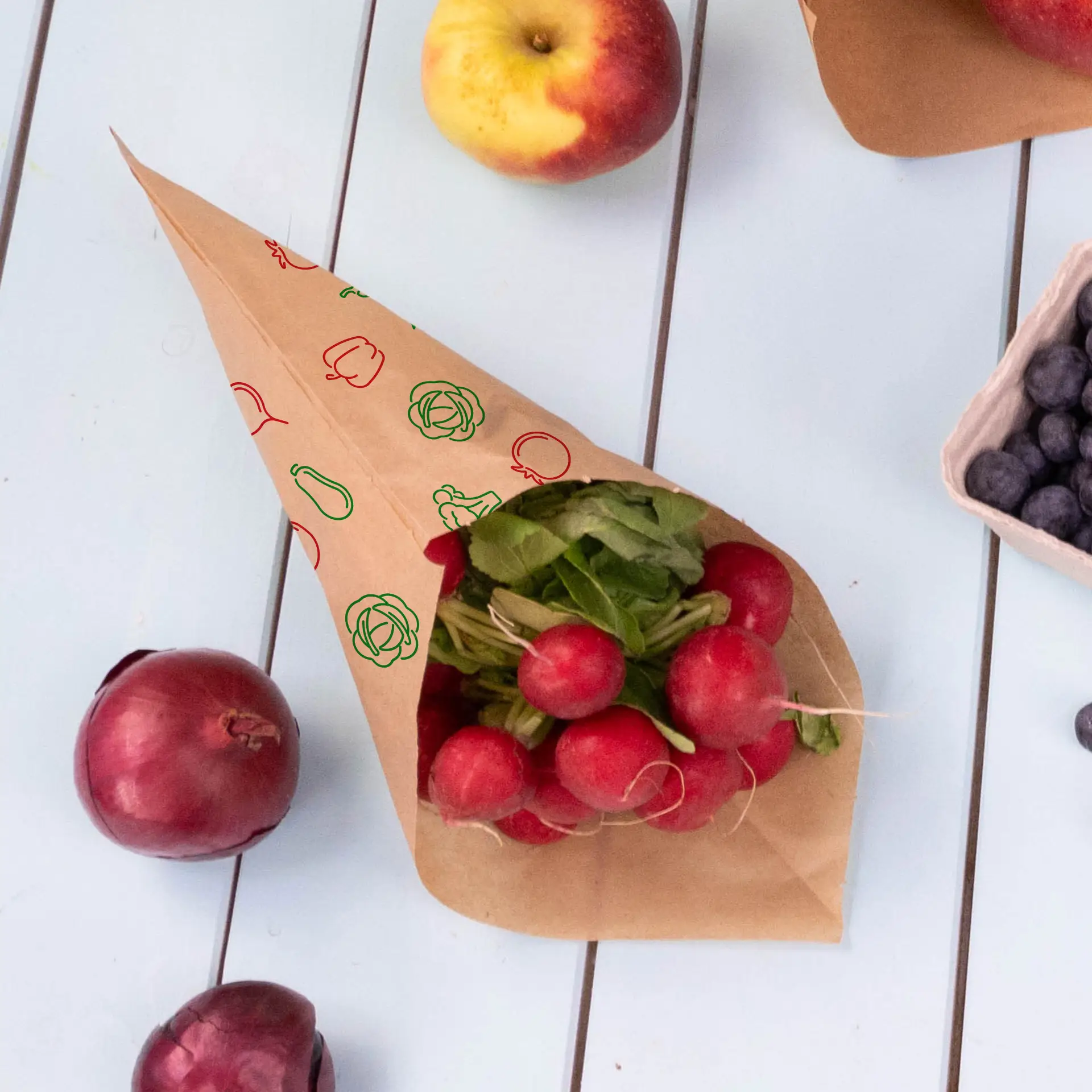 Cono di carta marrone riempito di ravanelli e erbe, circondato da mele, cipolle rosse e mirtilli su una superficie chiara.