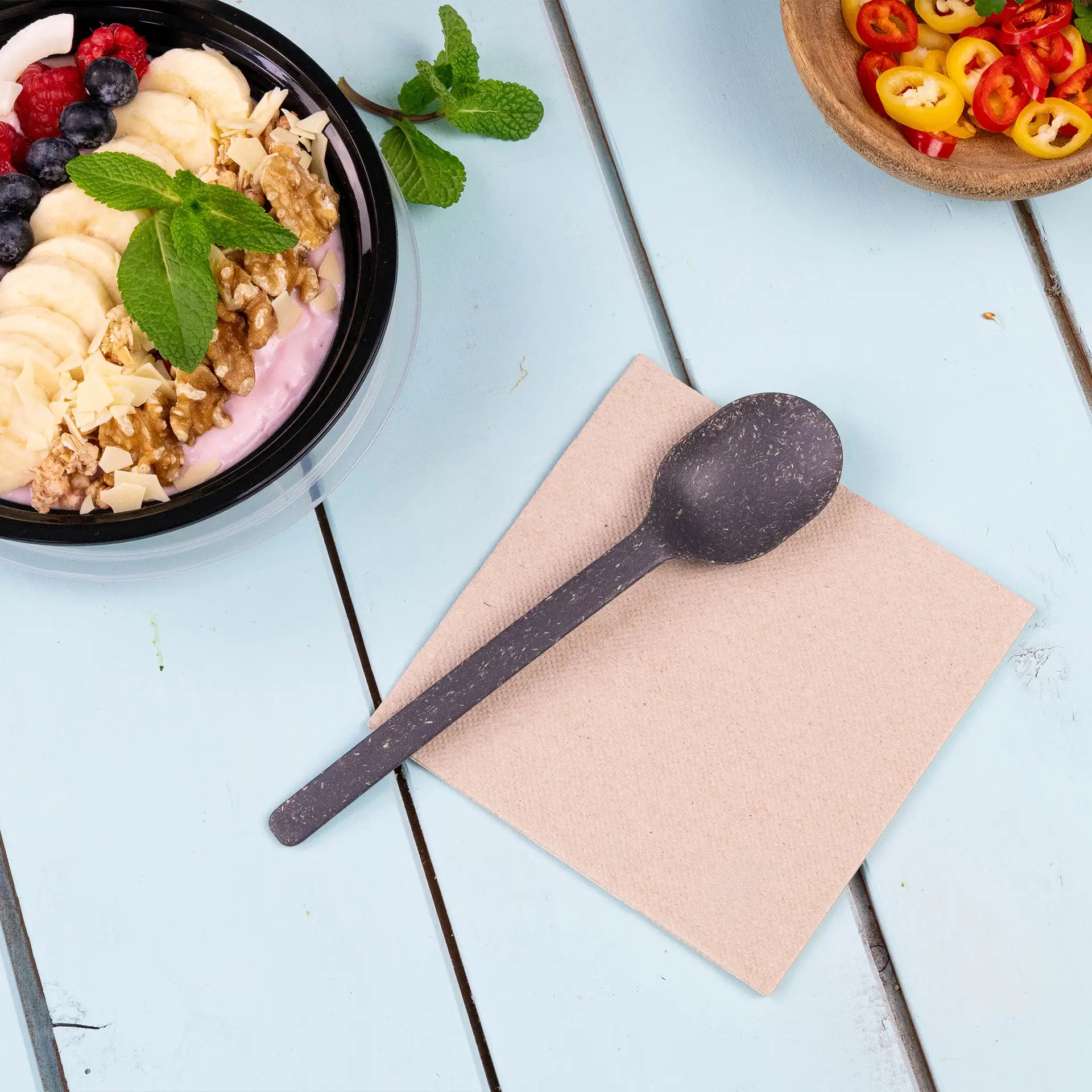 Image d'une cuillère noire sur une serviette beige, avec un bol de yaourt garni de fruits et de noix en arrière-plan.