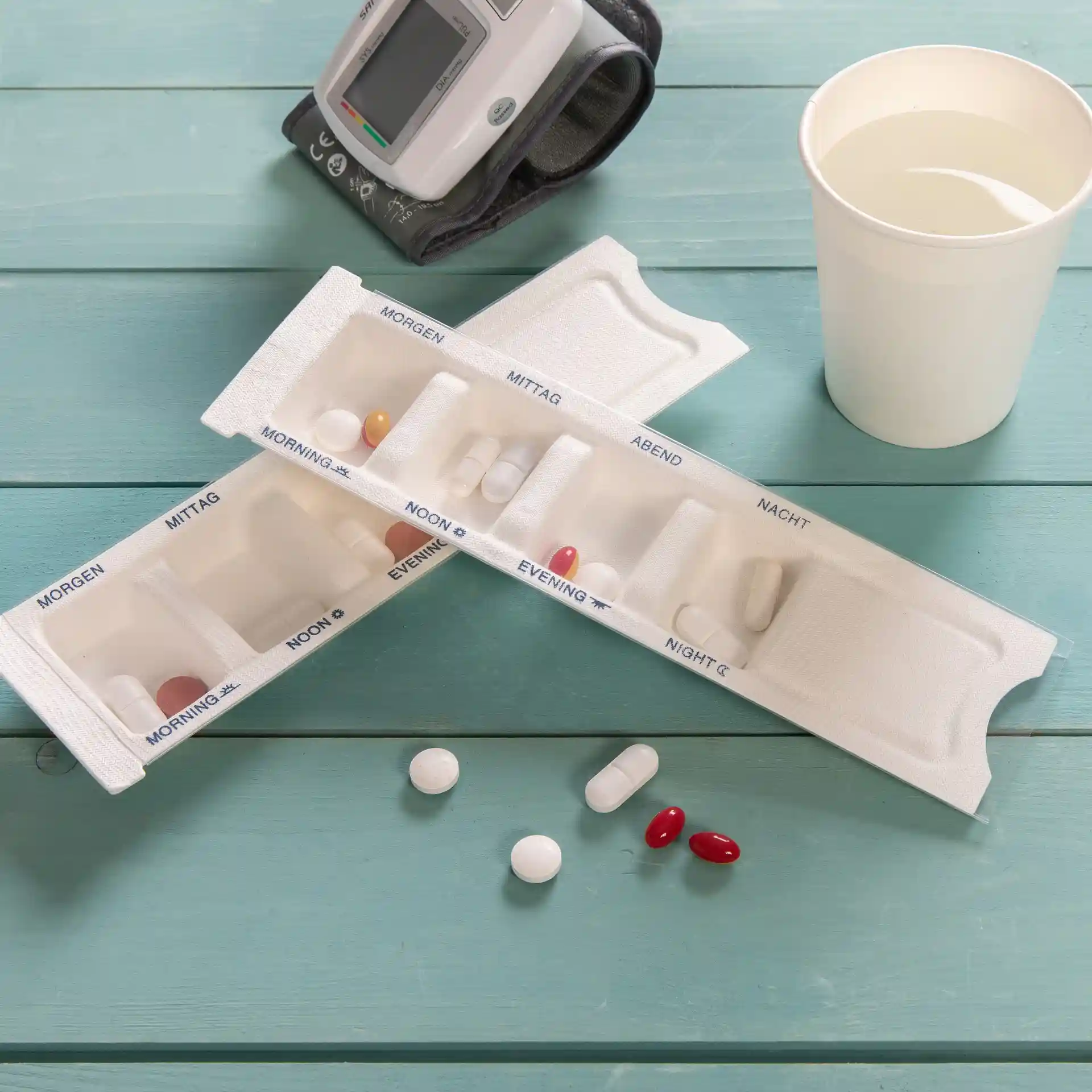 Distributeur de médicaments à quatre compartiments, couleur blanche, avec plusieurs pilules sur une surface bleue à côté d'un verre d'eau.